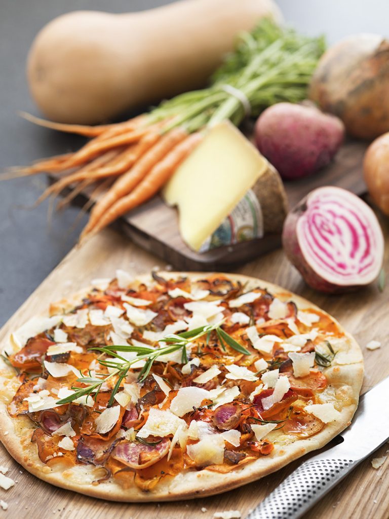 Pizza med färska grönsaker och ost på träskärbräda, bakverk och rotfrukter i bakgrund, fräsch och smakrik matbild.