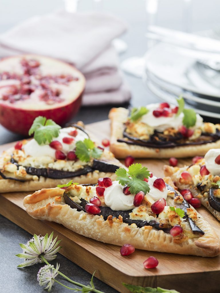 Färska pizzor med ricotta, aubergine och granatäpple på träbricka nära halverad granatäpple.